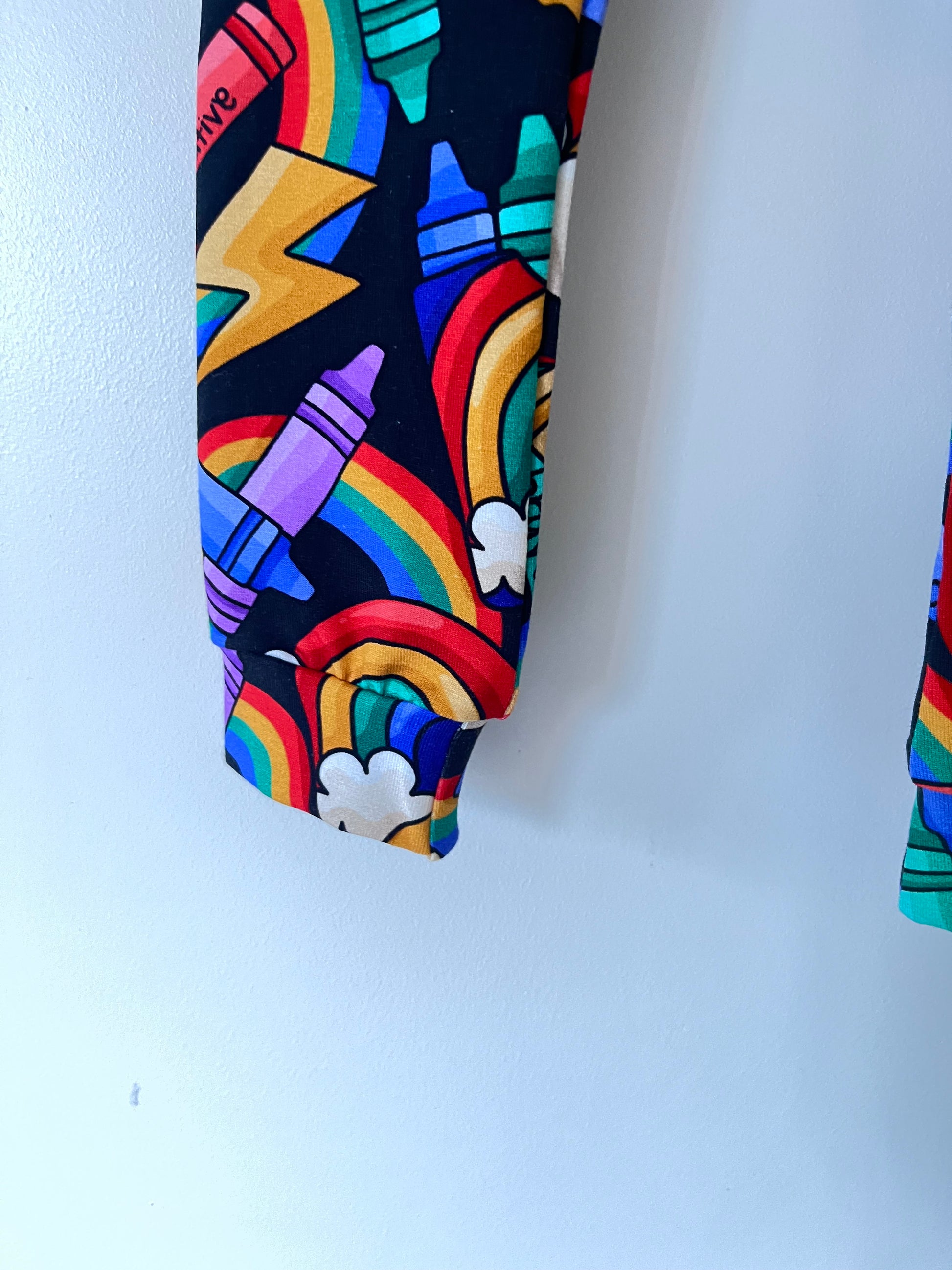 Handmade children’s leggings in a rainbow crayon print, featuring colourful crayons, rainbows, clouds, and positive messages on a dark background.