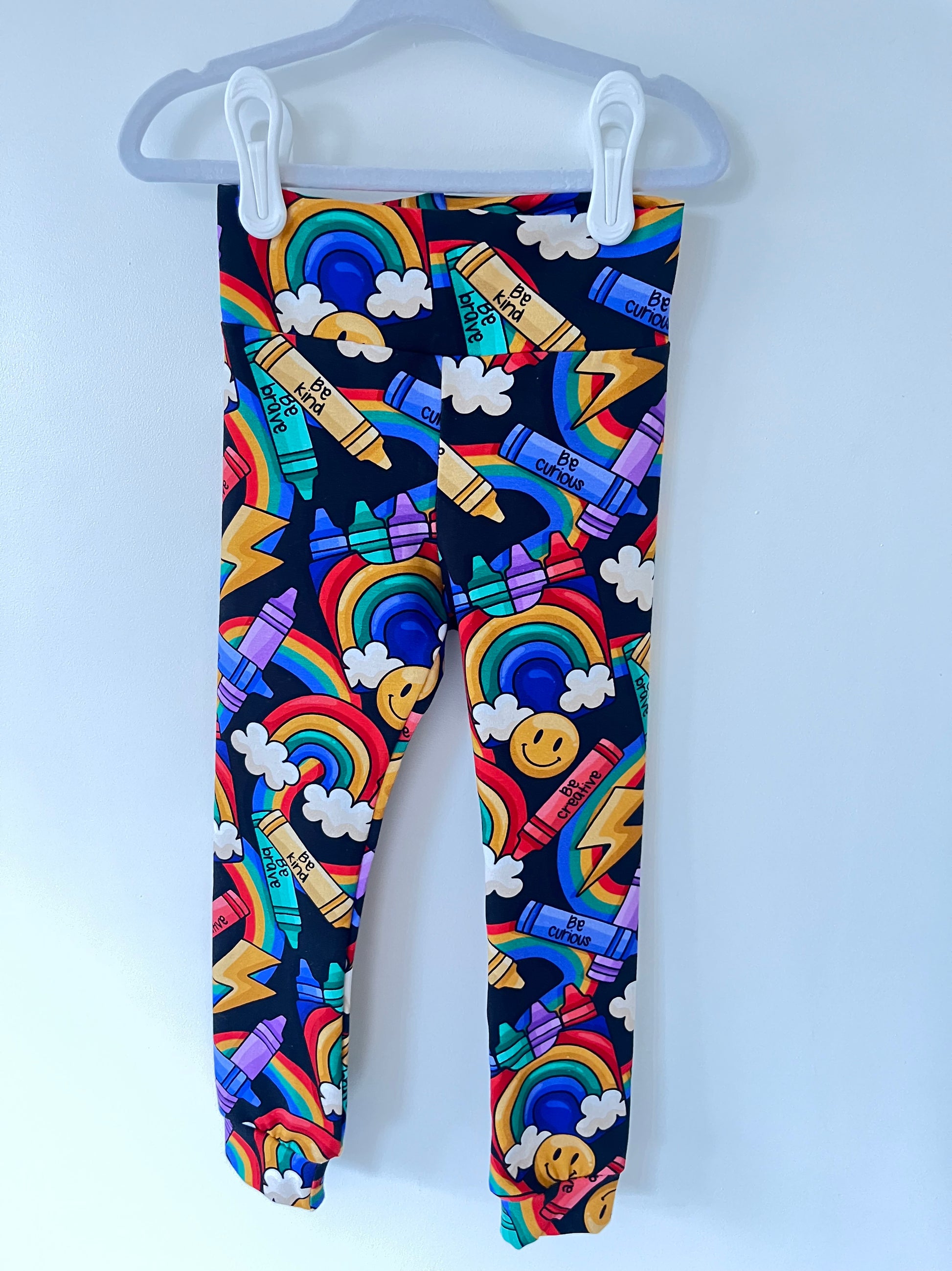 Handmade children’s leggings in a rainbow crayon print, featuring colourful crayons, rainbows, clouds, and positive messages on a dark background.